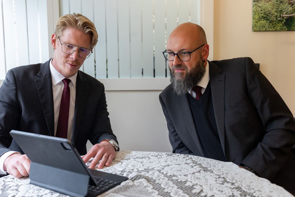 Zwei Männer in Anzügen schauen sich gemeinsam einen Laptop an einem Tisch in einem hellen Büro an.