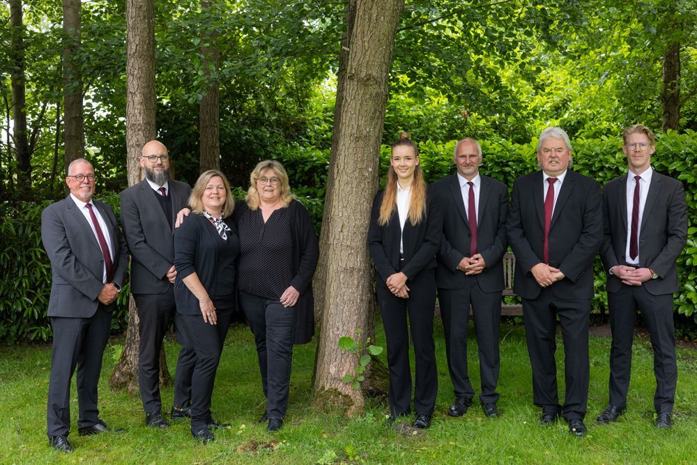Eine Gruppe von acht Personen in Anzügen steht im Freien auf einer Wiese neben einem Baum in einem Waldgebiet.