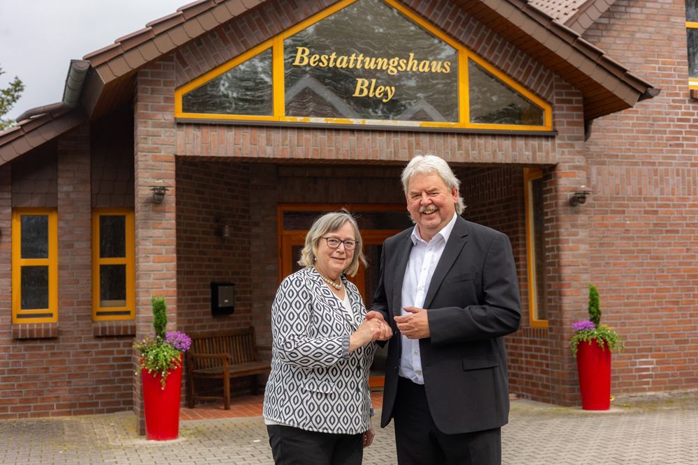Zwei Personen stehen vor einem Backsteingebäude mit einem Schild mit der Aufschrift „Bestattungshaus Bley“.