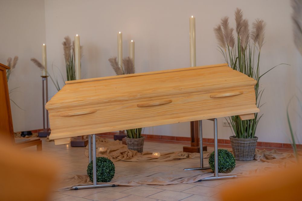 Ein hölzerner Sarg auf Ständern in einem kerzenbeleuchteten Raum mit Blumenarrangements.