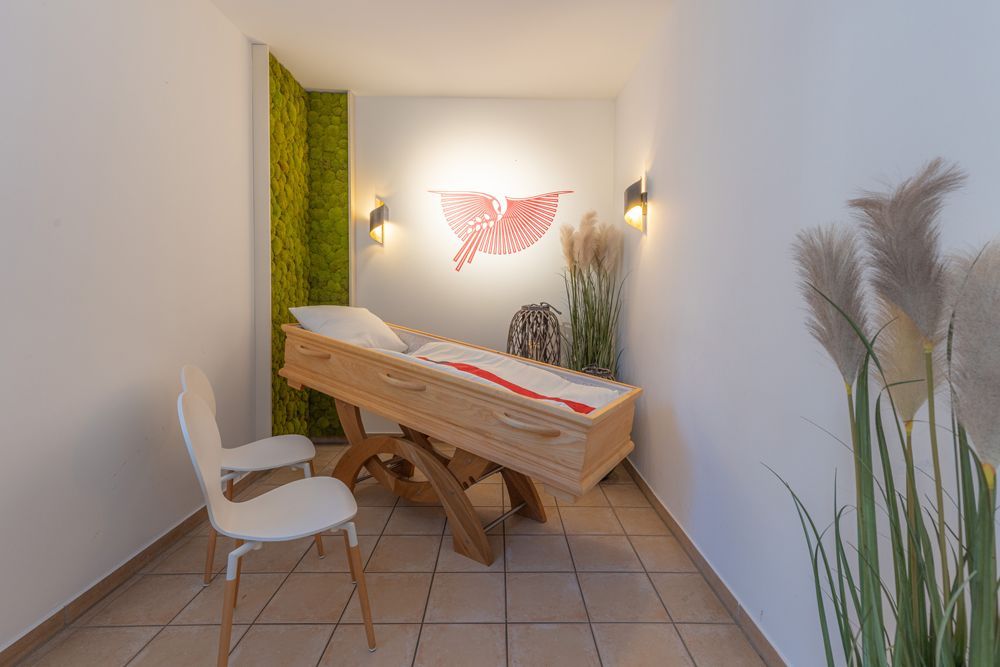 Helles, kleines Zimmer mit einer Holzbank, weißen Stühlen, Wandbildern und hohen Pflanzen