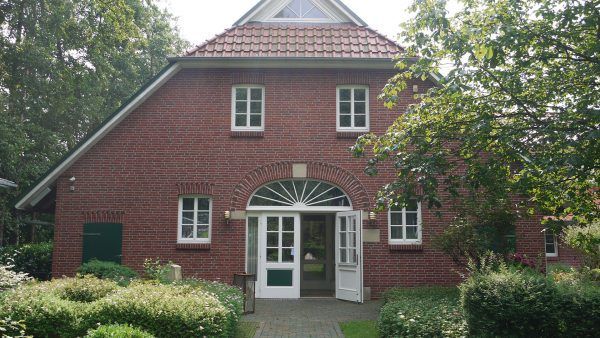 Backsteinhaus mit weiß umrandeten Fenstern und gewölbter Eingangstür, umrahmt von Bäumen und Sträuchern.