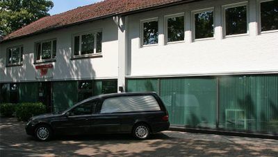Ein schwarzes Auto parkte vor einem zweistöckigen weißen Gebäude mit grünen Fenstern und einem roten Ziegeldach.