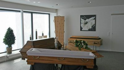 Ein spärlich eingerichteter Raum mit gestapelten Särgen oder Tischen, einem Topfbaum und Wandbildern an großen Fenstern