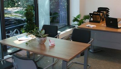 Modernes Büro mit Schreibtischen, Stühlen, einem Laptop, Papieren und einer Pflanze an großen Fenstern.