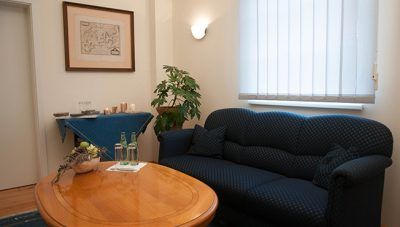 Gemütliches Wohnzimmer mit marineblauem Sofa, rundem Holztisch, blauem, stoffbezogenem Beistelltisch, Pflanze und Wandbild.