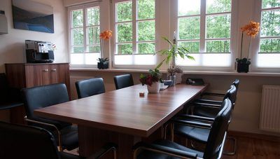 Konferenzraum mit Holztisch, schwarzen Stühlen, großen Fenstern und Topfblumen auf der Fensterbank