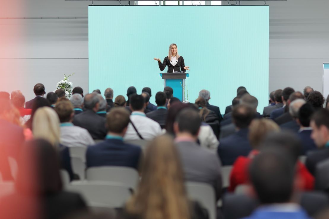 Una mujer está dando un discurso a un grupo grande de personas en una sala de conferencias.