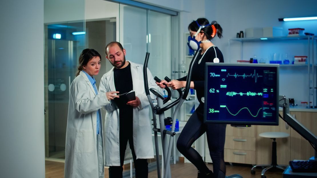 Una mujer está montando una bicicleta estática en un laboratorio mientras dos médicos la observan.