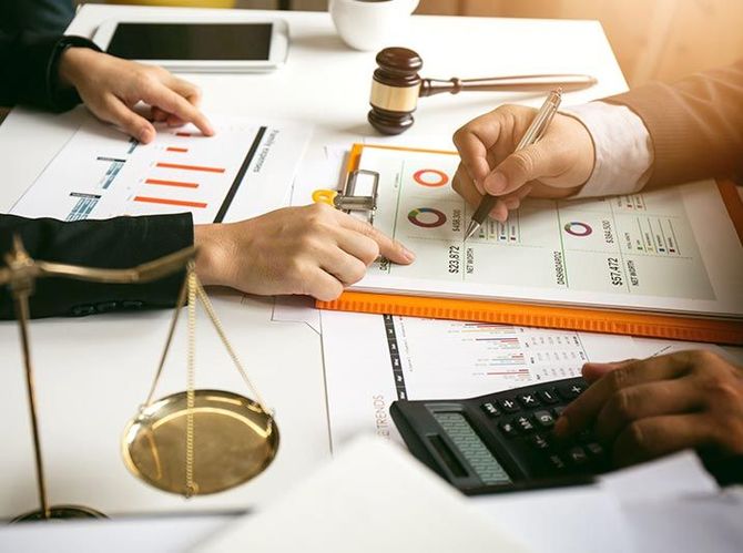 Dos personas revisan documentos financieros con una calculadora, un mazo y una balanza de la justicia sobre un escritorio.