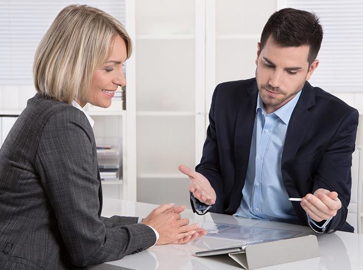 Una mujer y un hombre de traje, sentados en un escritorio, revisan documentos. El hombre gesticula con un bolígrafo, la mujer sonríe en la oficina.