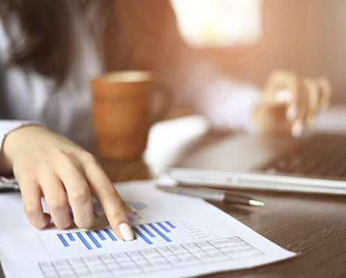 Mujer señalando un gráfico de barras en un documento, mientras trabaja en una computadora portátil con una taza de café cerca.