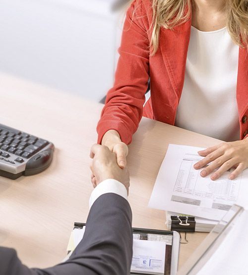 Dos personas se dan la mano en un escritorio. Una mujer con chaqueta roja y papeles cerca.
