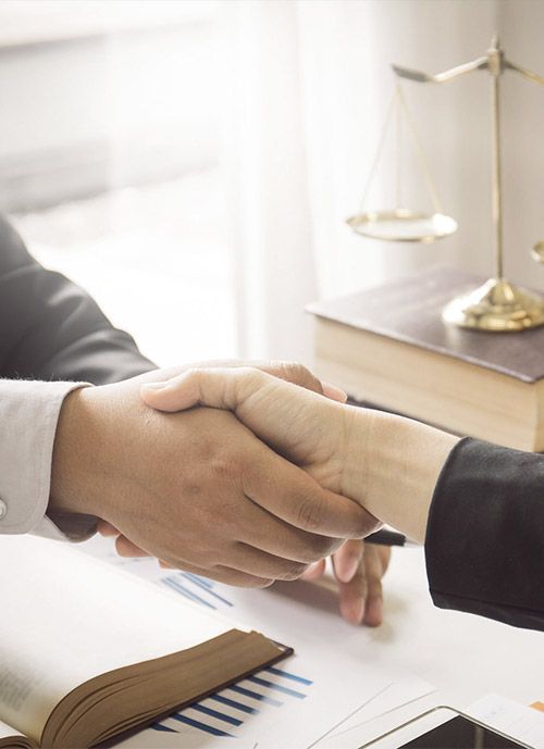 Dos personas se dan la mano en un escritorio, con una balanza de justicia y libros de leyes en el fondo.