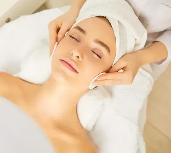 Mujer recibiendo un tratamiento facial; acostada con los ojos cerrados, toallas, manos limpiando la cara con algodones.
