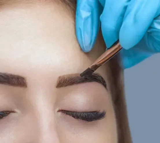 Persona que se tiñe las cejas: la mano enguantada del técnico aplica el tinte con una brocha. Ojos cerrados, primer plano.