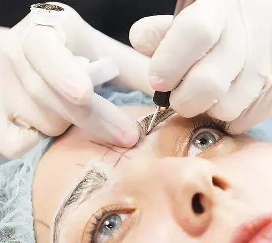 Persona que se realiza microblading de cejas; técnico con guantes sosteniendo una herramienta sobre la ceja marcada.