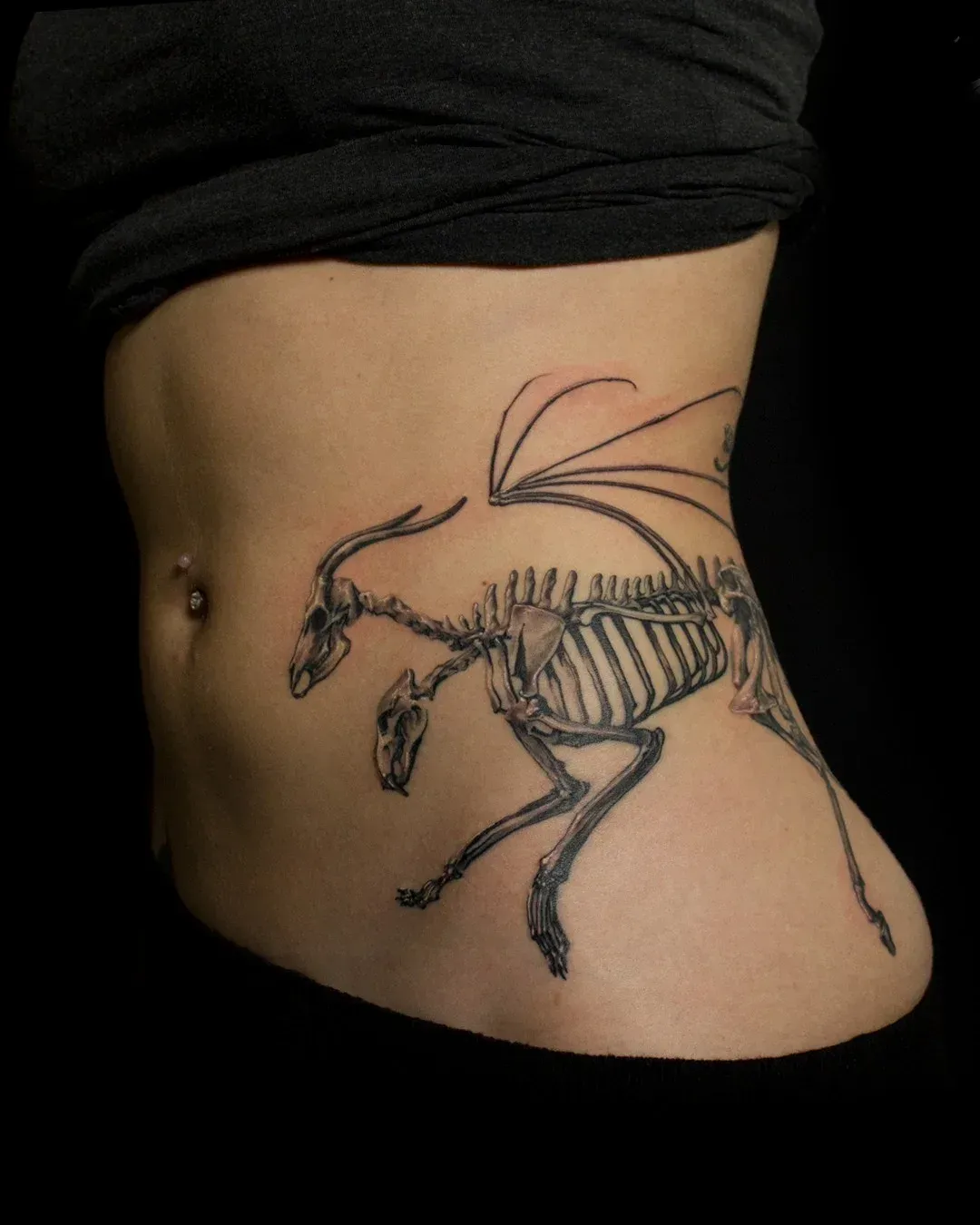 Tatuaje de un caballo alado esquelético en el costado de una persona, ombligo visible, camisa negra.