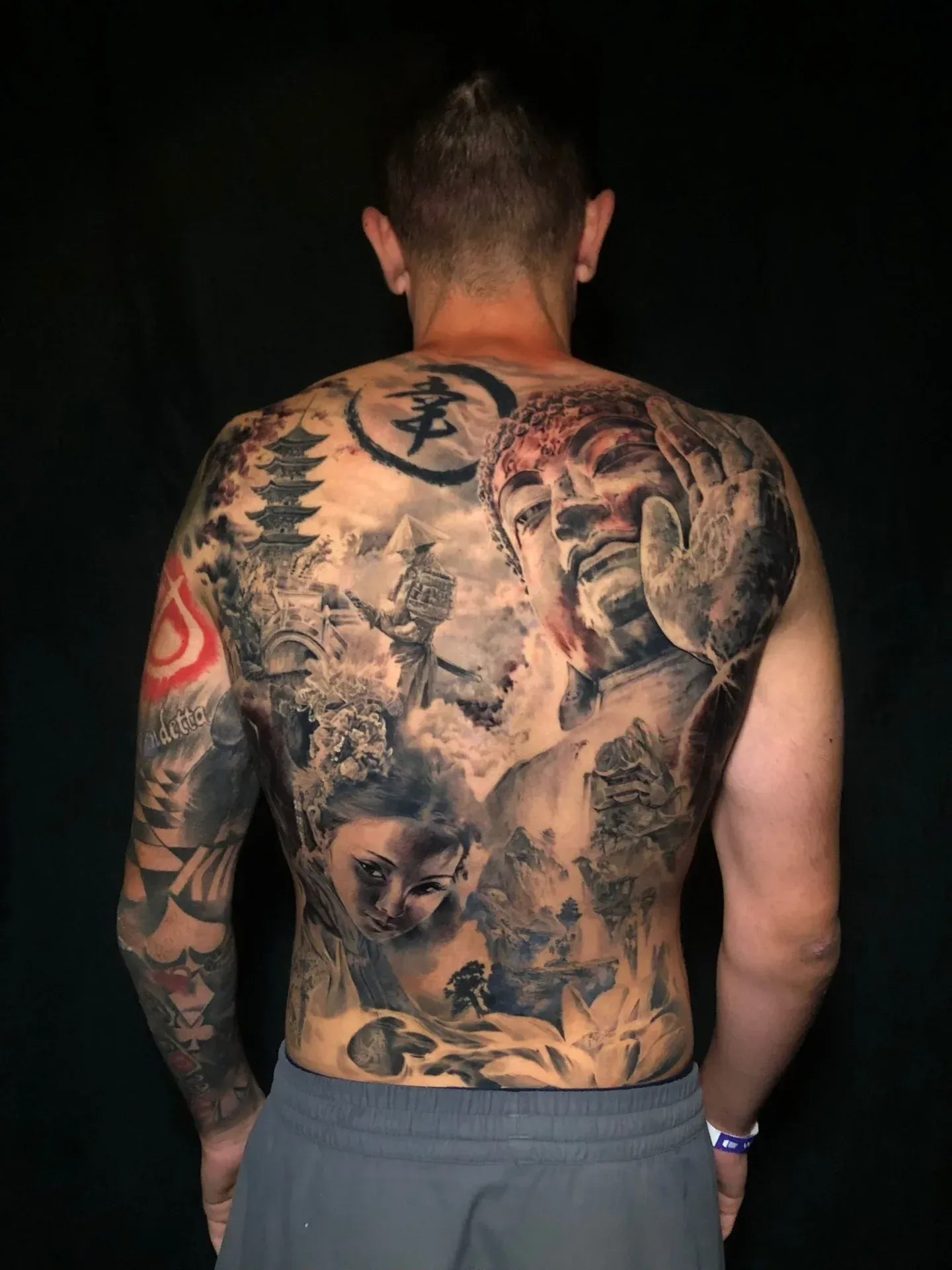 Tatuaje en la espalda con imágenes de inspiración asiática, que incluyen un Buda, pagodas y paisajes, en negro y gris.
