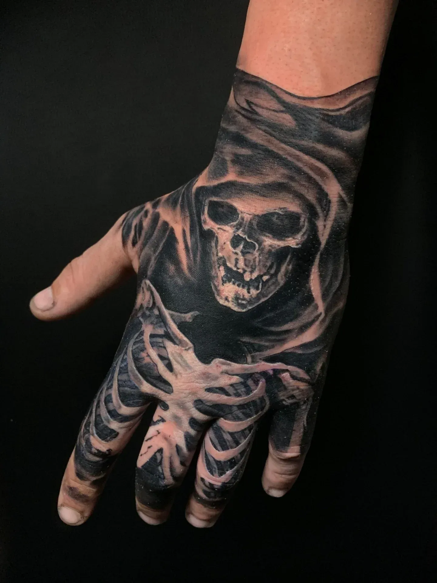 Tatuaje de calavera y mano esquelética de la Parca en negro y gris sobre un fondo oscuro.