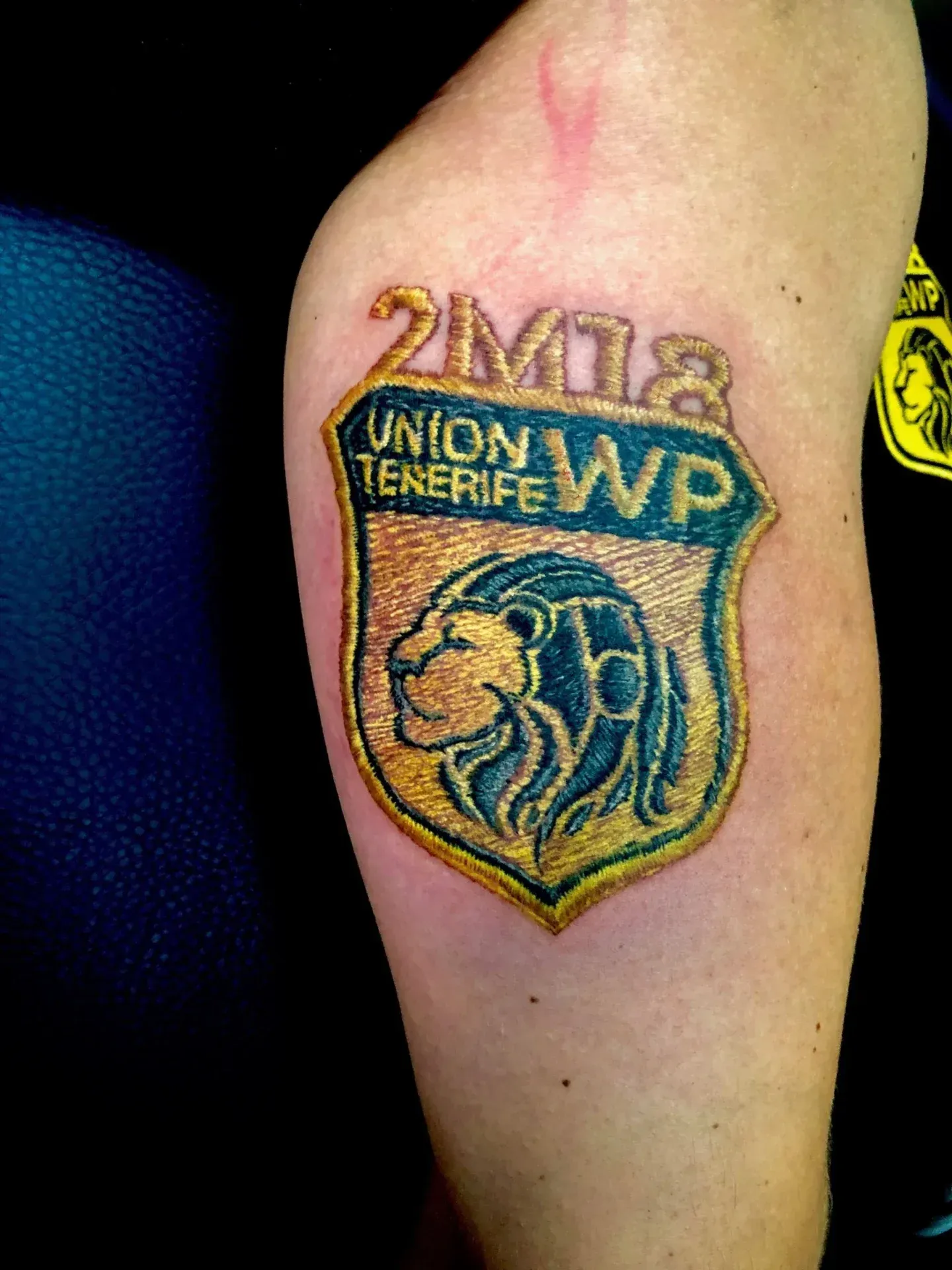 Tatuaje del logo de Unión Tenerife en el brazo de una persona, dorado, azul, con cabeza de león y el año 2M18.