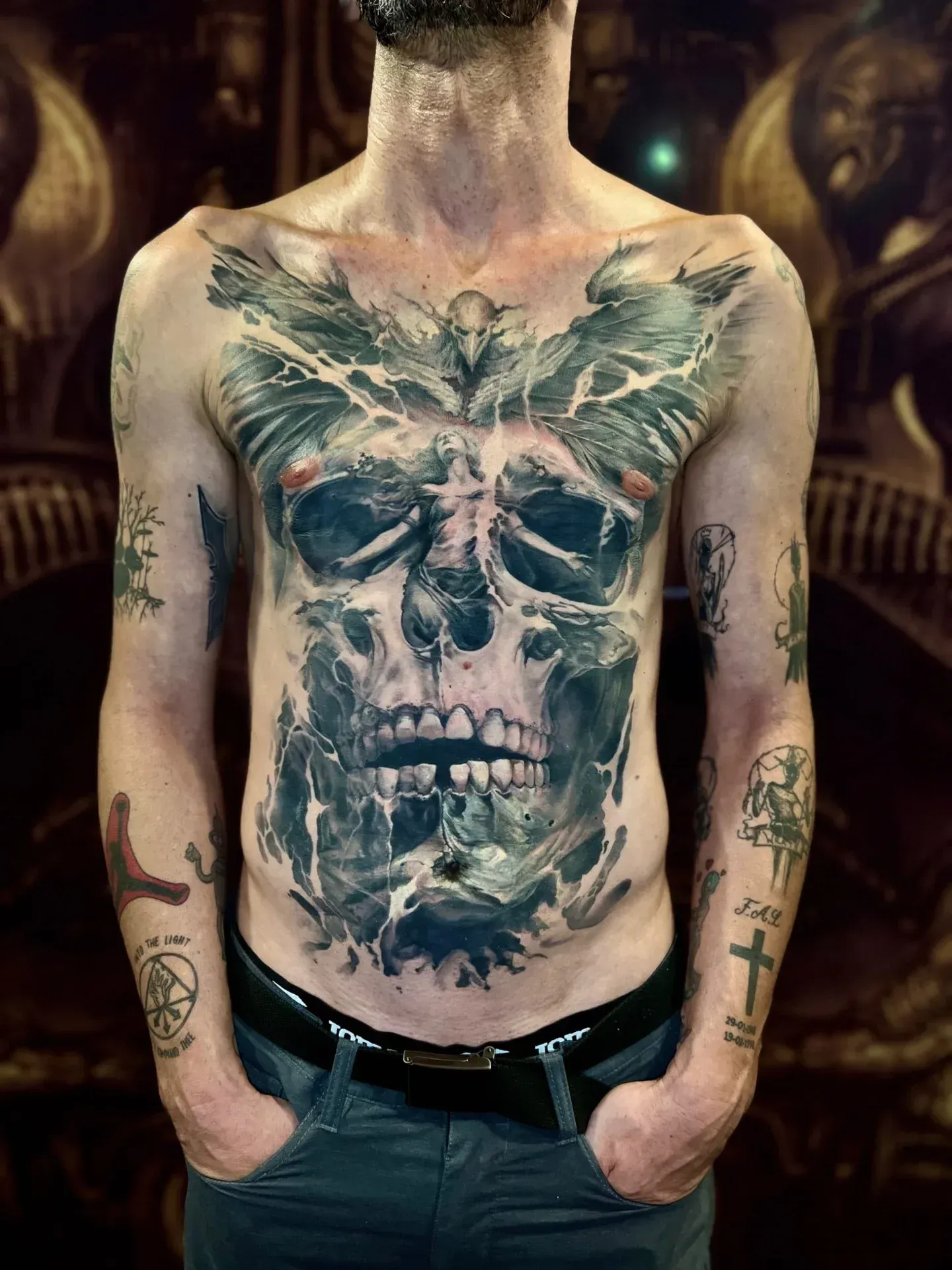 Hombre con un gran tatuaje de calavera en el torso, con las manos en los bolsillos. El tatuaje es negro y gris.