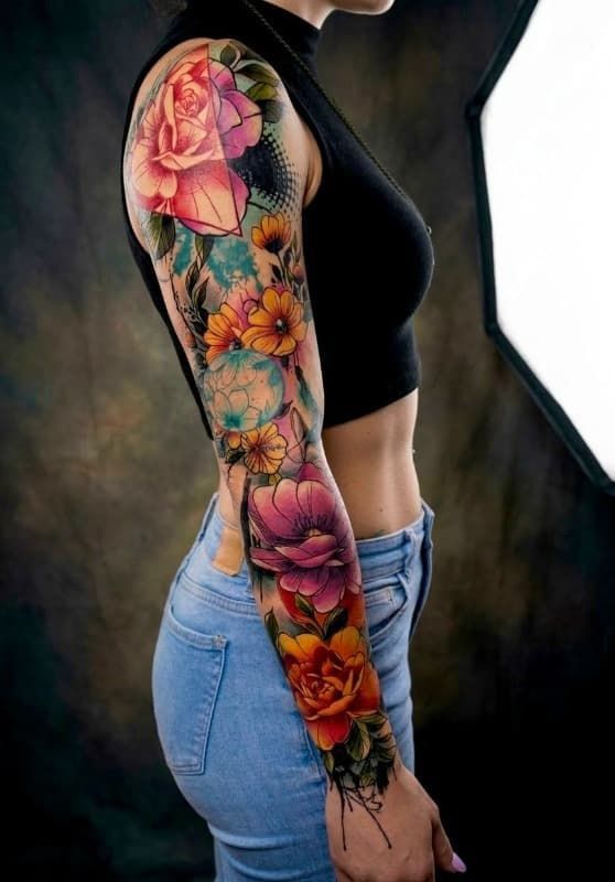Vista lateral de un brazo tatuado con flores de colores, que lleva una blusa negra y vaqueros azul claro sobre un fondo oscuro.