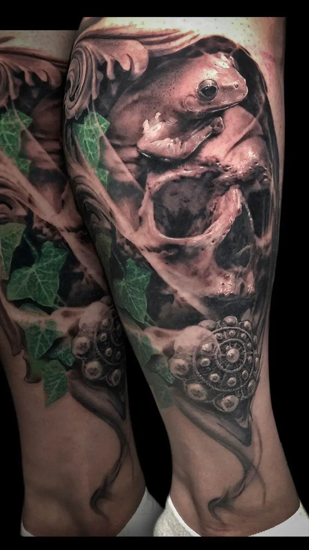 Tatuaje de una calavera con una rana en su interior, hiedra y detalles ornamentales en blanco y negro con acentos verdes.