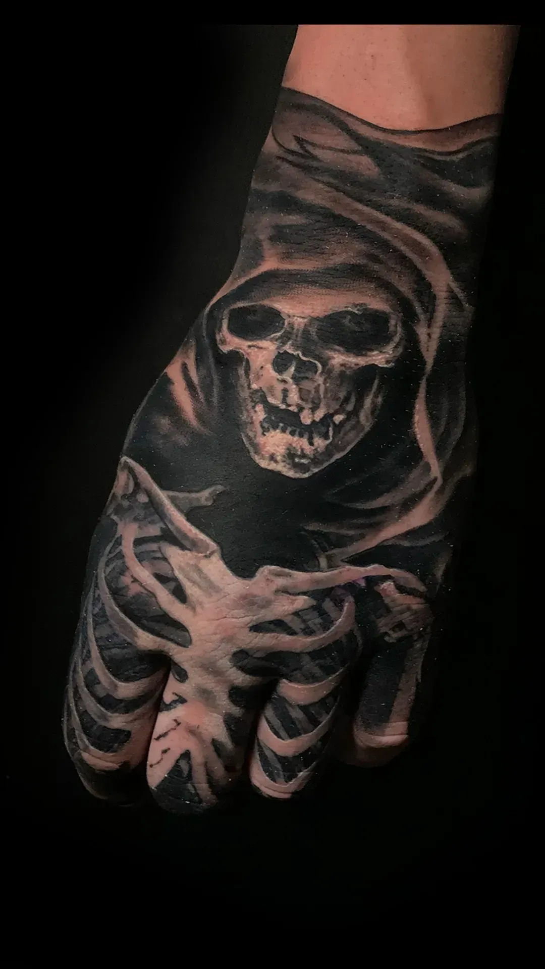 Tatuaje en la mano de una calavera y caja torácica, la calavera con capucha oscura. Sombreado en negro y gris.
