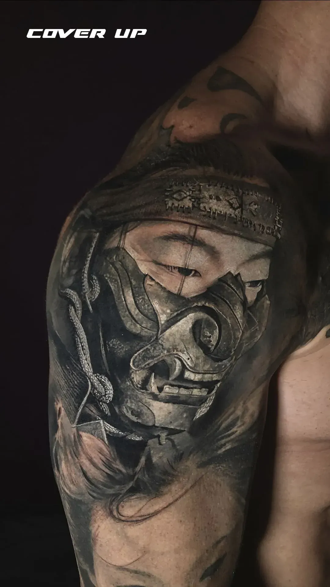 Tatuaje de una máscara rota en un hombro. Sombreado negro y gris con grietas visibles. Texto