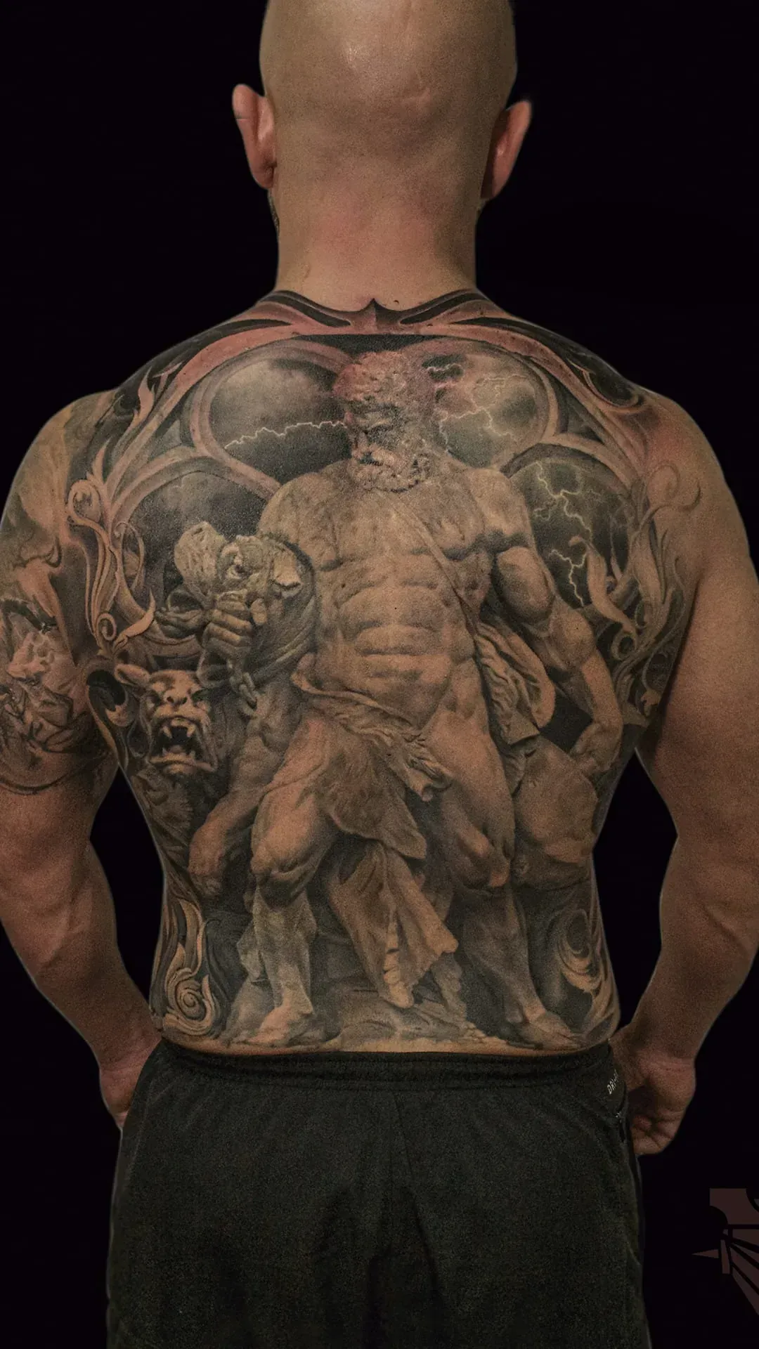 Tatuaje en toda la espalda: persona musculosa, figura masculina clásica, diseño intrincado en escala de grises, fondo negro.