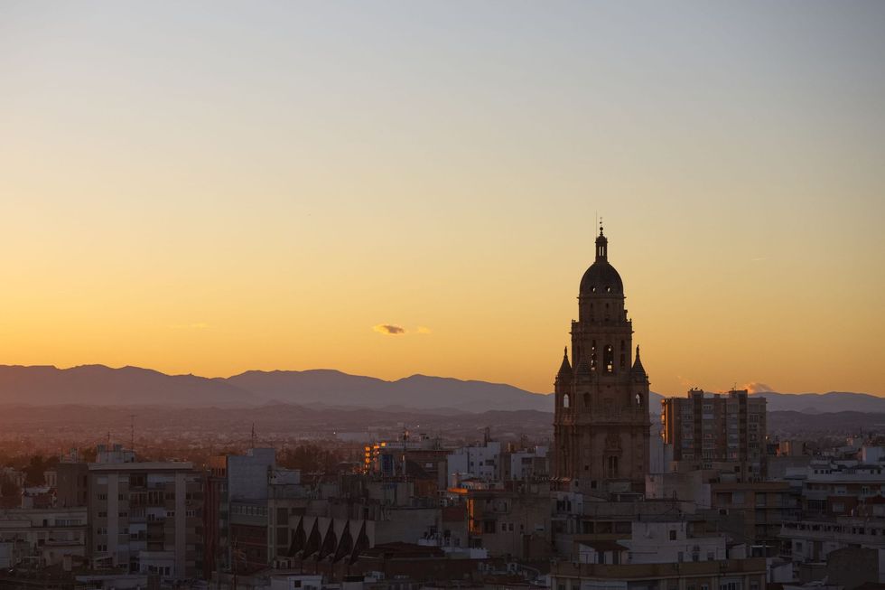 Horizonte urbano al atardecer con una alta torre histórica recortada contra un cielo dorado y montañas distantes.