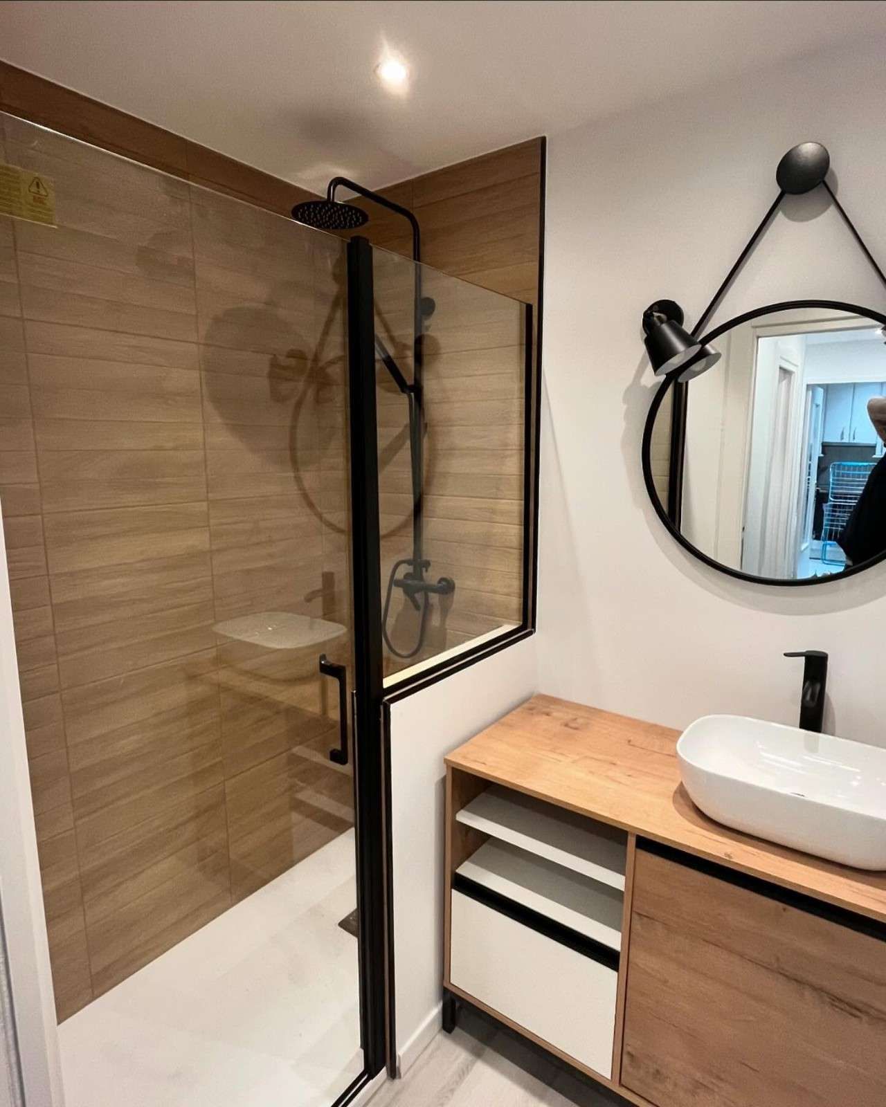 Baño moderno con ducha con apariencia de madera, accesorios negros y un tocador con espejo redondo.