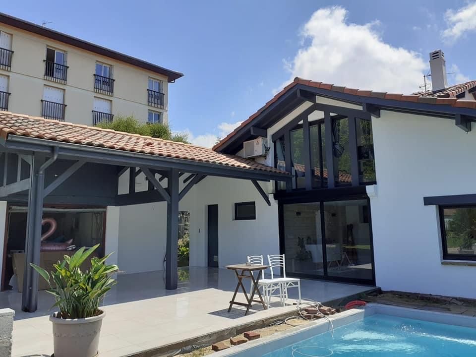 Moderna casa blanca con piscina, patio y edificio de apartamentos adyacente, bajo un cielo soleado.