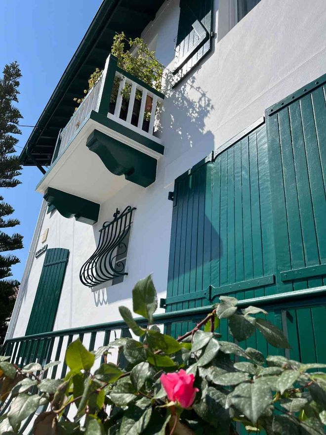 Casa blanca con persianas verdes y balcón, rosal en primer plano.