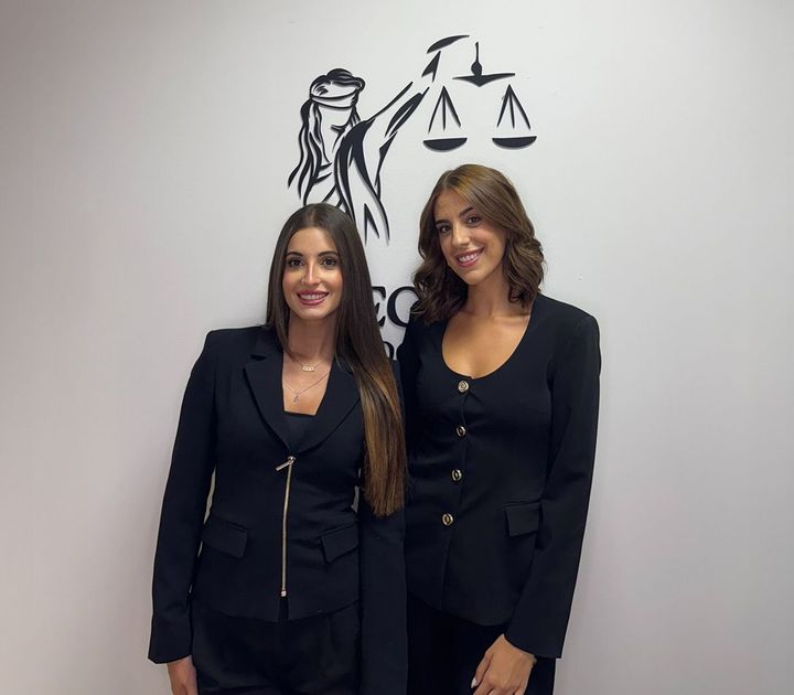 Dos mujeres vestidas con trajes negros posan sonrientes frente a un mural con temática legal.