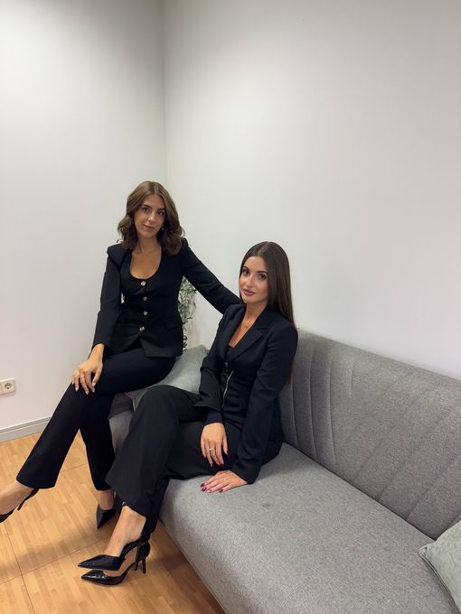 Dos mujeres vestidas de negro sentadas en un sofá gris en una habitación blanca.