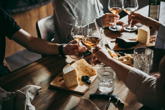 Menschen stossen mit Weingläsern an einem Holztisch mit Käse und Snacks an.