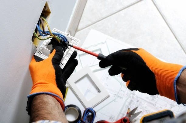 Electricista con guantes naranjas, instalando cableado en una pared, utilizando un destornillador.