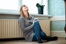 Una mujer está sentada en el suelo junto a un radiador sosteniendo una taza de café.