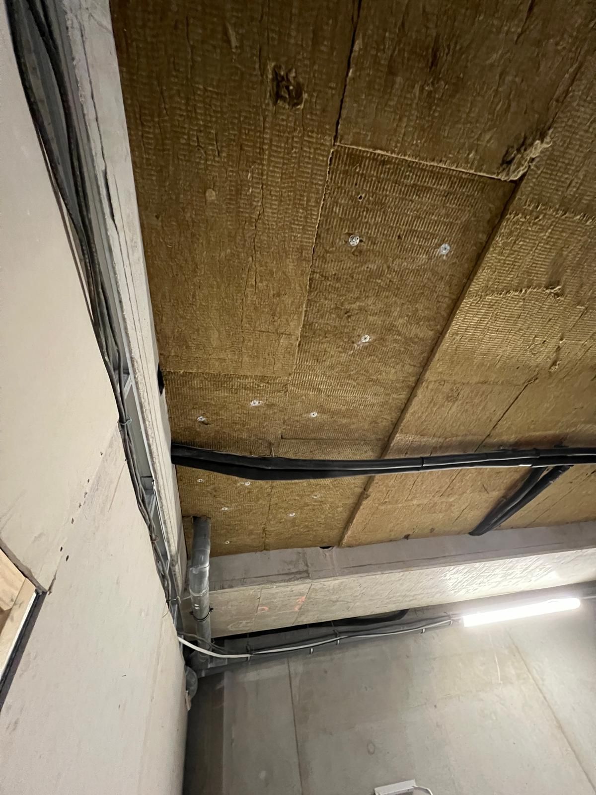 Isolation posée au plafond. Câbles noirs et supports métalliques apparents. Isolation beige, murs blancs et gris.