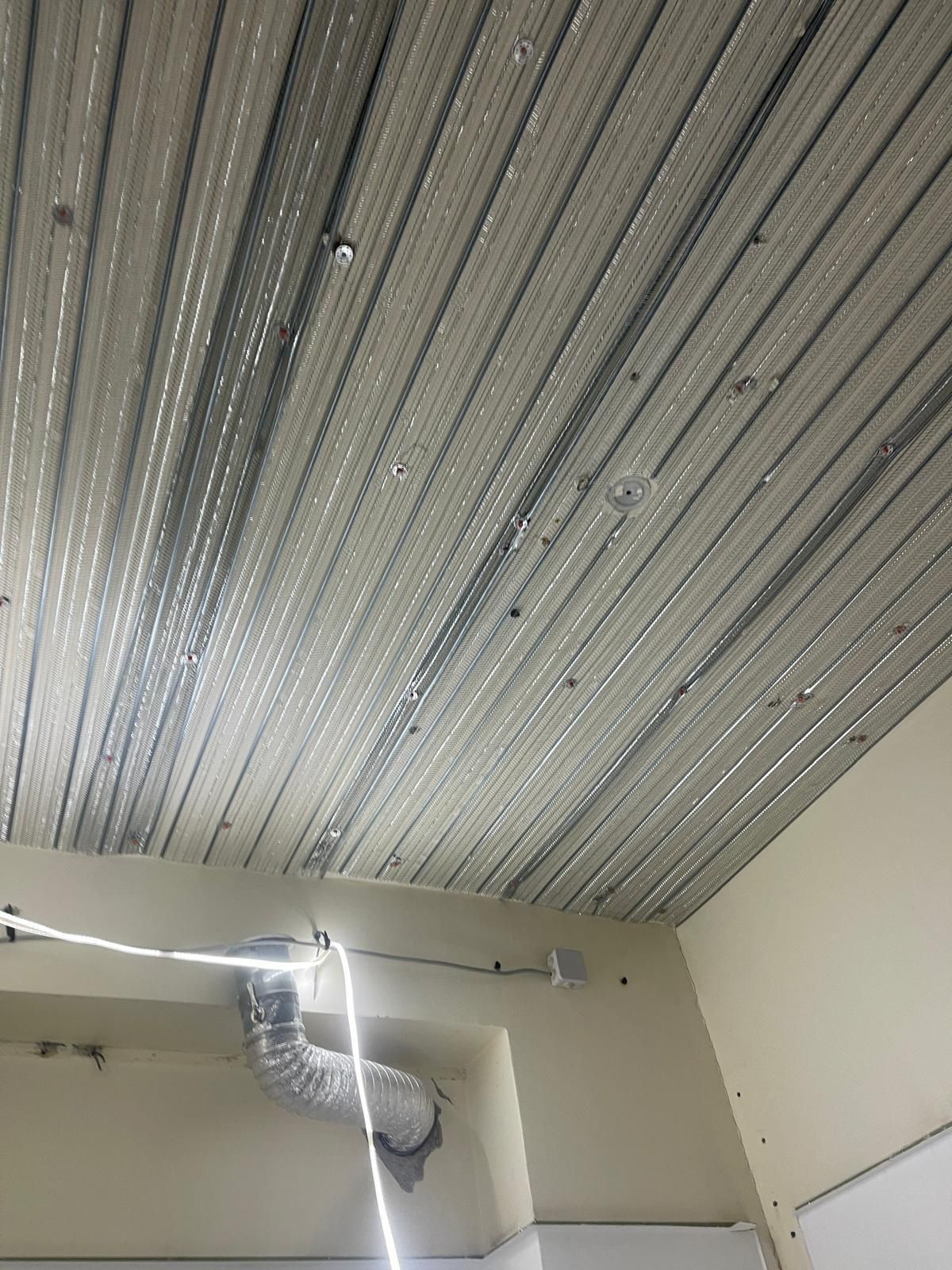 Plafond avec barres métalliques et grille d'aération, murs couleur crème et luminaire blanc.