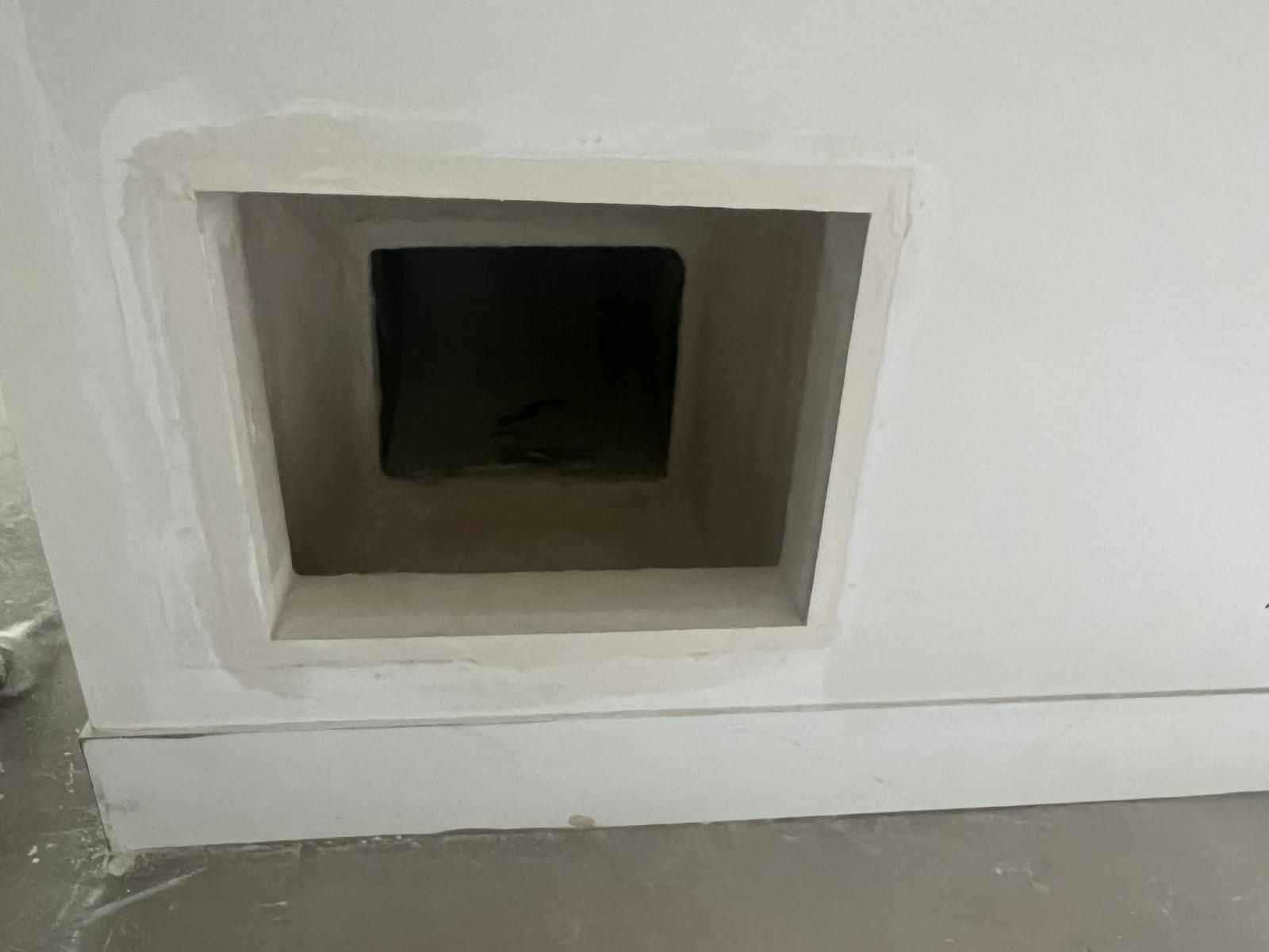 Une ouverture carrée en retrait dans un mur blanc, avec une bordure en plâtre et un intérieur sombre.