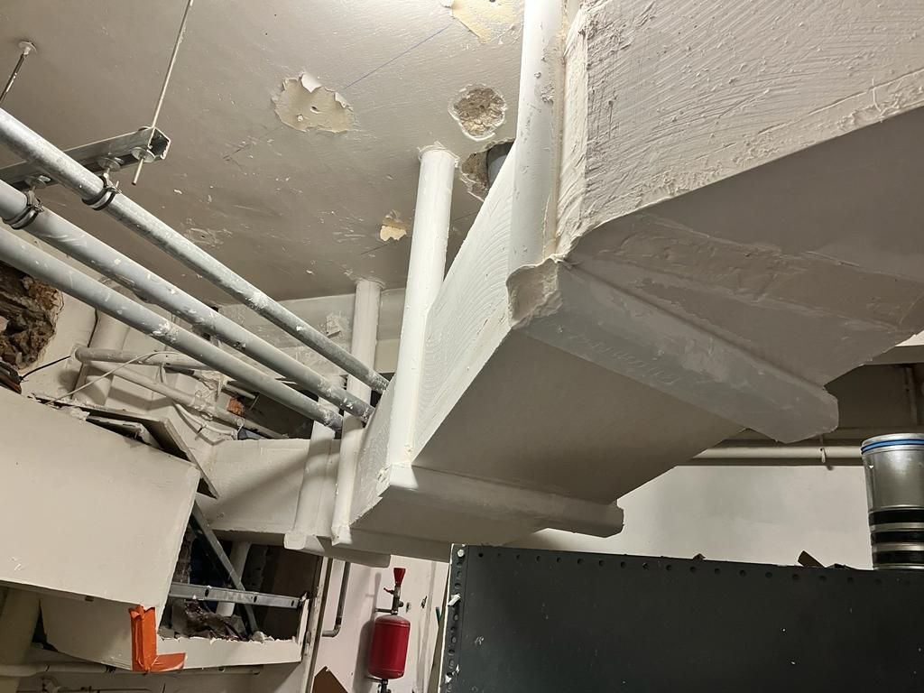 Plafond blanc endommagé, laissant apparaître des tuyaux, une poutre et un extincteur.