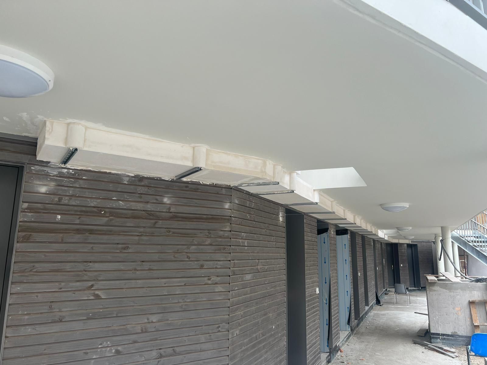 Vue extérieure d'un bâtiment en construction, montrant une passerelle avec un plafond en béton et un mur en bois.