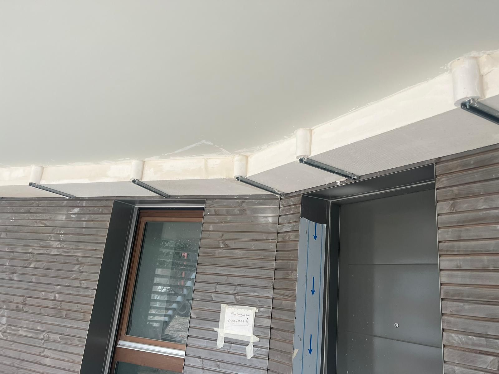 Extérieur d'un bâtiment en construction ; façade en bois gris, fenêtre, supports métalliques le long d'un plafond blanc.