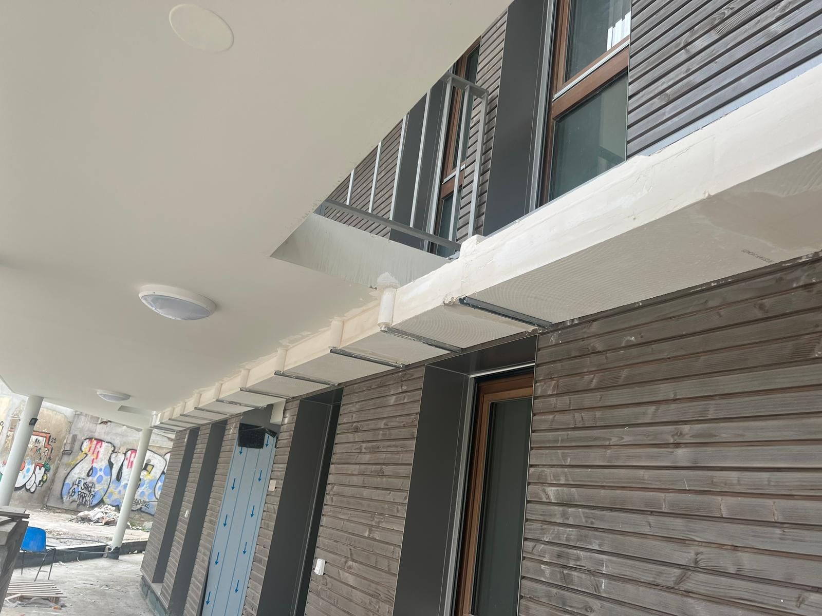 Vue extérieure d'un bâtiment avec un bardage en bois, un plafond blanc et un balcon en métal.