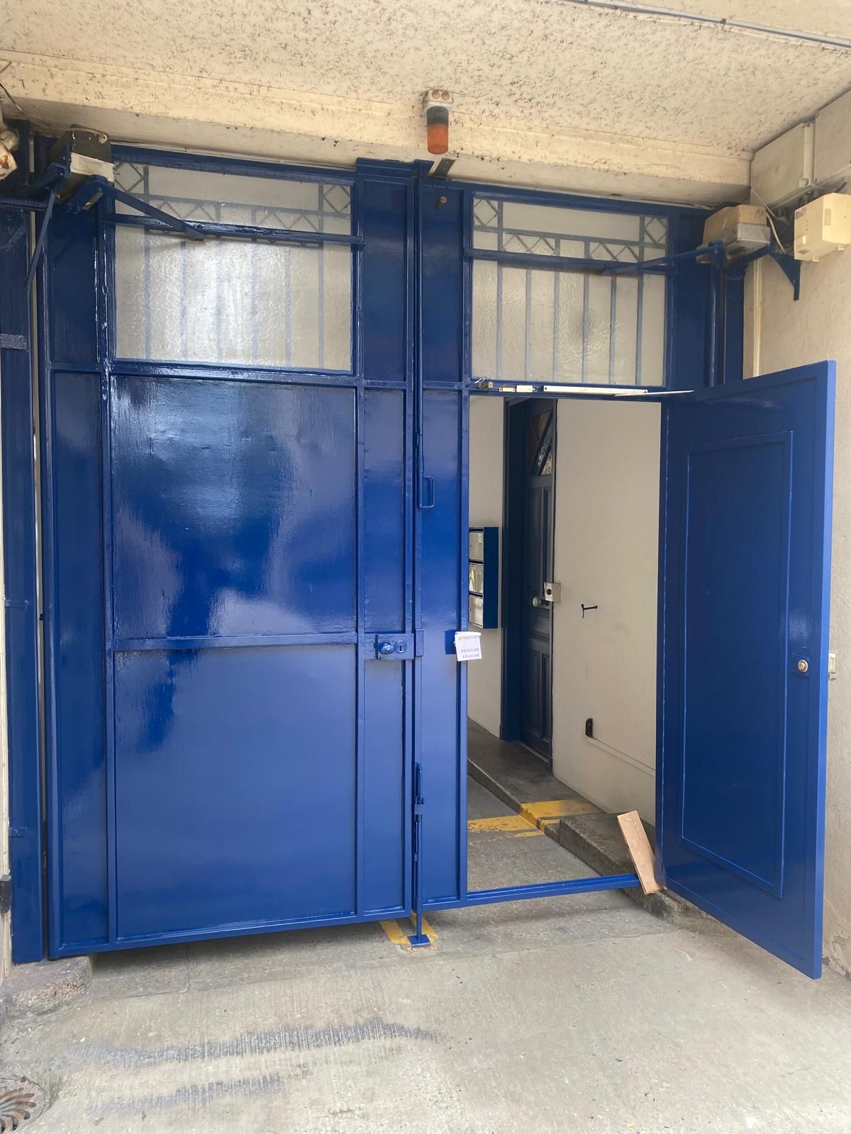 Entrée en béton avec des portes métalliques bleues, dont une entrouverte, qui donnent sur un bâtiment.