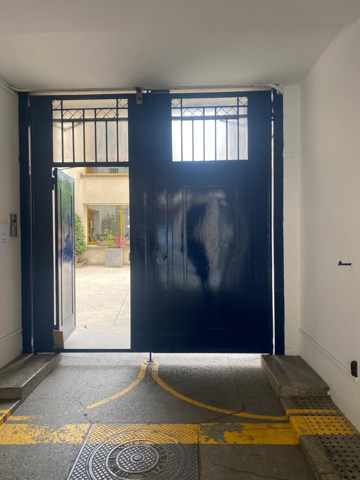 Un portail en métal, entrouvert, donne sur une cour intérieure. Des lignes jaunes au sol.