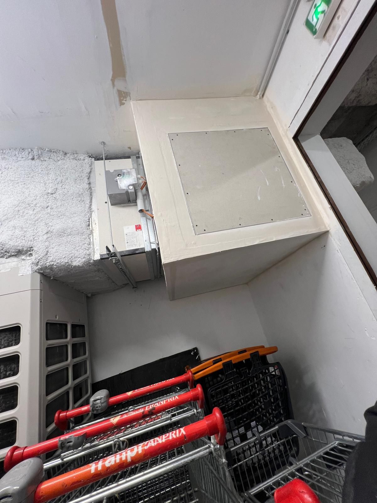 Des chariots de supermarché empilés sous un système de ventilation fixé au plafond et muni d'une trappe d'accès.